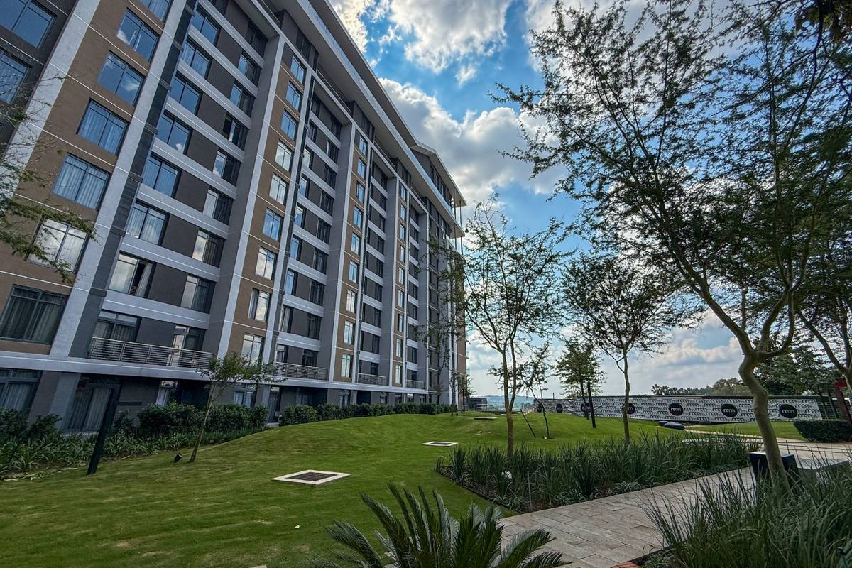 A view of the apartment building exterior featuring landscaped gardens, paved walkways, and modern architectural design with multiple balconies.