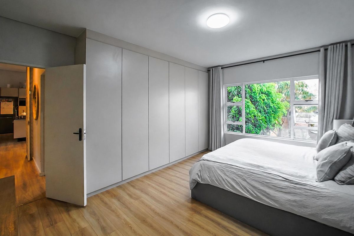 A well-maintained bedroom featuring extensive built-in cupboards, modern laminate flooring, and large windows providing natural light and garden views.