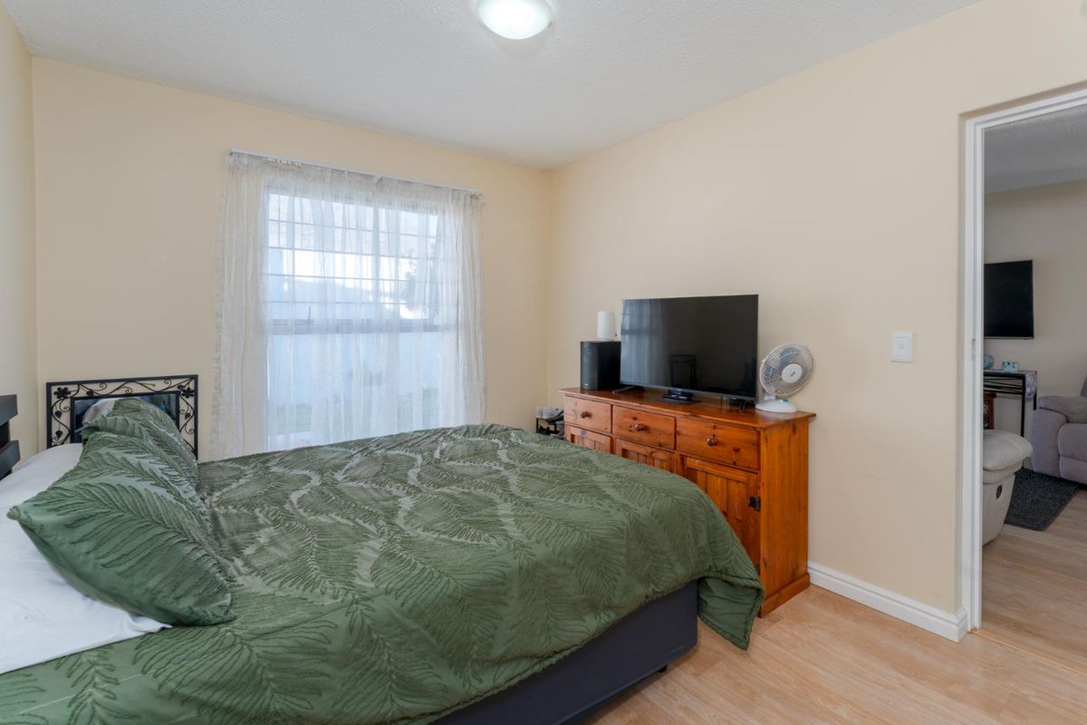 A functional bedroom featuring light-toned laminate flooring, a window with sheer curtains, and a doorway leading to an adjacent space.
