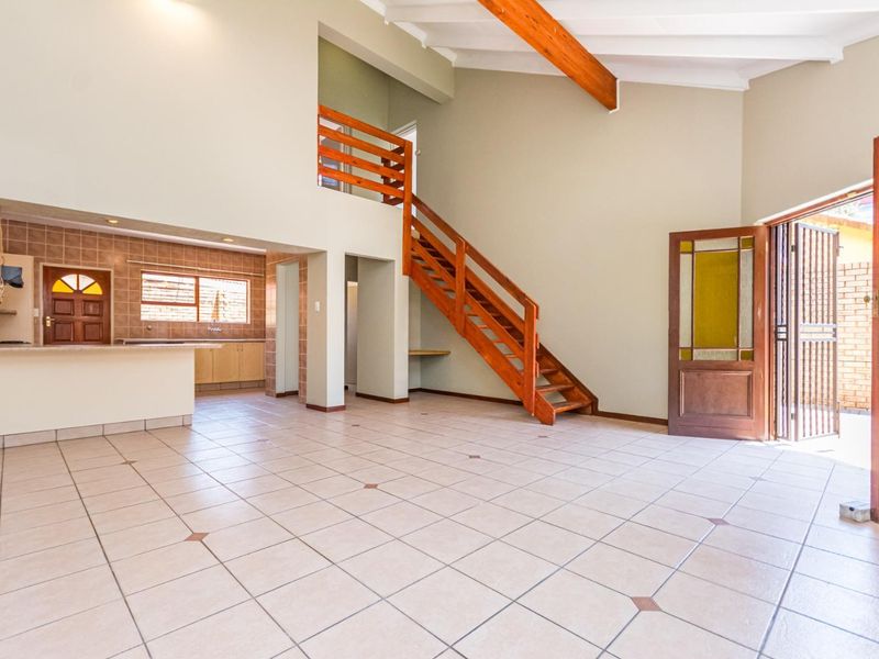 Spacious open-plan living area featuring a double-volume ceiling with exposed beams, a wooden staircase, and an adjoining kitchen with tiled finishes.