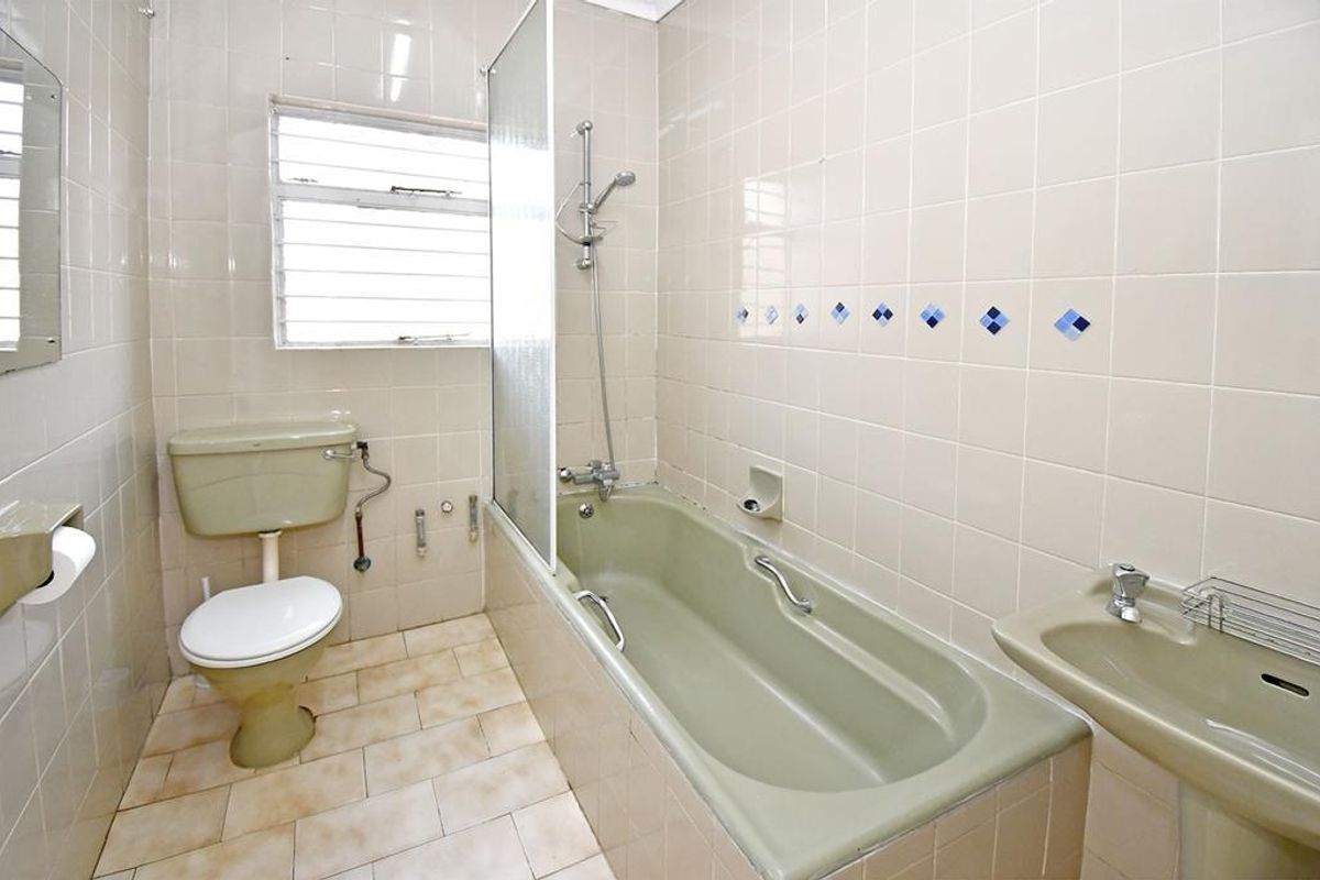 A dated bathroom featuring a vintage olive-toned bathtub, pedestal basin, and toilet, complemented by wall-to-wall tiling and a glass shower screen.