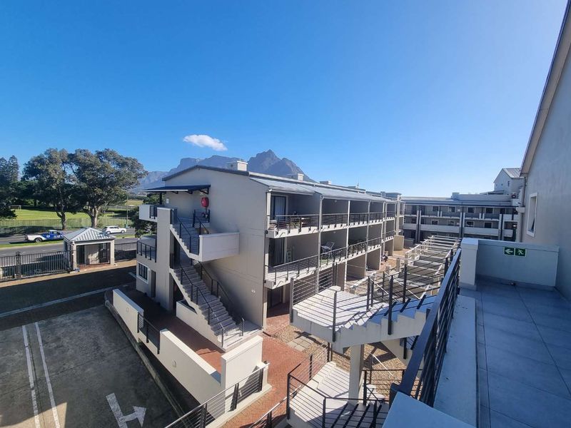 An elevated view of the apartment complex exterior, featuring multi-level balconies, external staircases, paved walkways, and mountain views in the background.