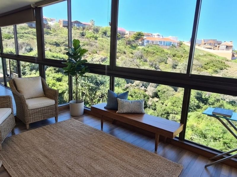 A bright, enclosed balcony featuring expansive glass windows that offer panoramic views of the surrounding hillside and natural landscape.