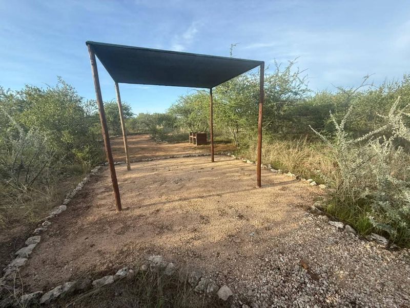 An outdoor gravel patio area featuring a simple metal shade structure, set within a natural bushveld landscape.