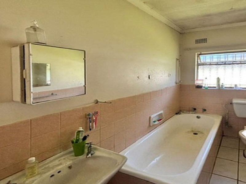 A dated bathroom featuring a standard bathtub, pedestal basin, and toilet, with tiled splashbacks and a window with security bars.