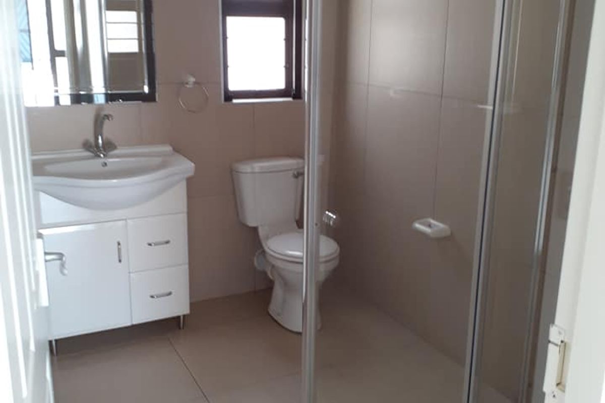 A clean, modern bathroom featuring a glass-enclosed shower, a white vanity with a basin, and a standard toilet with neutral floor-to-ceiling tiling.