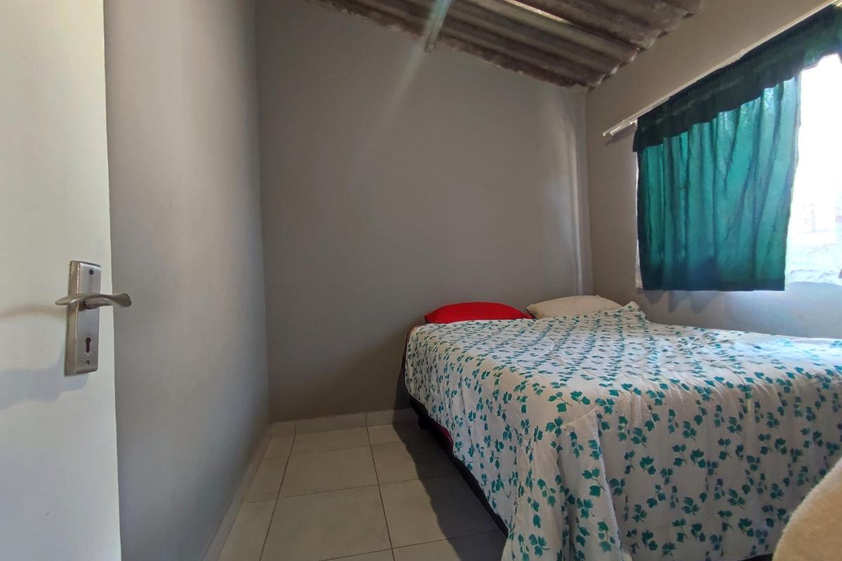 A compact bedroom featuring tiled flooring and an exposed corrugated roof structure, providing a basic and functional living space.