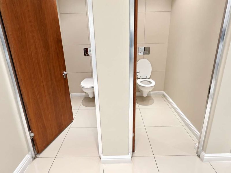 A clean, modern office bathroom facility featuring two private stalls with wall-mounted toilets, tiled flooring, and wood-finish doors.