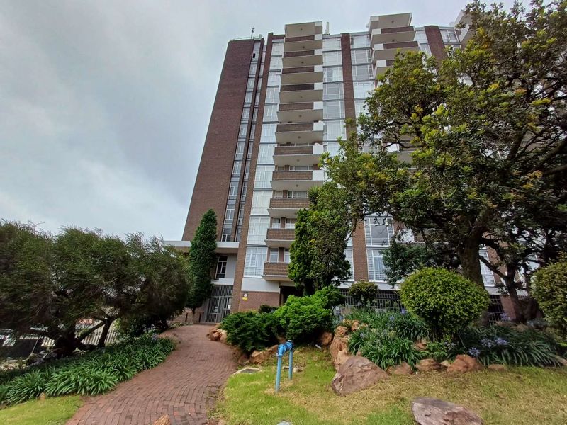 Exterior view of a multi-story apartment building featuring a brick facade, private balconies, and a landscaped garden with a paved walkway.