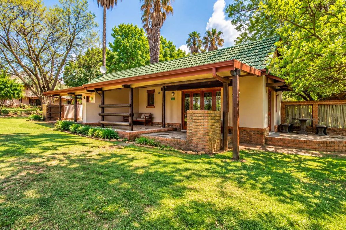 A well-maintained lodge exterior featuring a covered patio with brick pillars, timber beams, and a tiled roof, set within a lush garden.