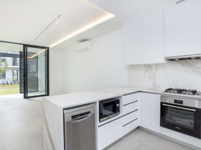Modern open-plan kitchen featuring sleek white cabinetry, stone countertops, integrated appliances, and direct access to a private garden area through sliding glass doors.