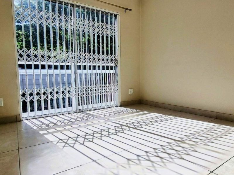 A bright, empty living area featuring neutral tiled flooring and a sliding glass door secured with a metal security gate.