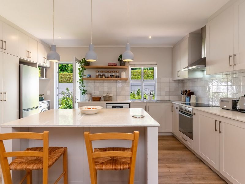 Modern, well-lit kitchen featuring a central island, quartz countertops, built-in oven, extractor fan, and stylish floating shelves with direct access to the garden.