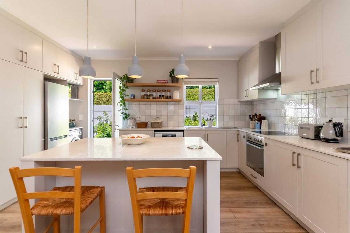 Modern, well-lit kitchen featuring a central island, quartz countertops, built-in oven, extractor fan, and stylish floating shelves with direct access to the garden.