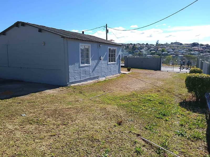 Rear view of a single-story concrete block house with a metal roof, security window bars, and a fenced yard area.
