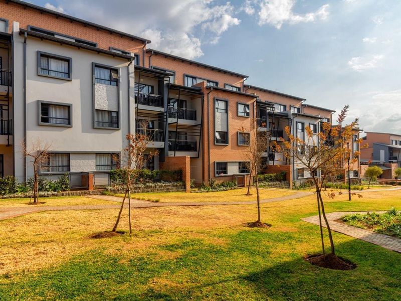 A view of the apartment complex rear facade featuring brick and plaster finishes, private balconies, and well-maintained communal garden areas with paved pathways.