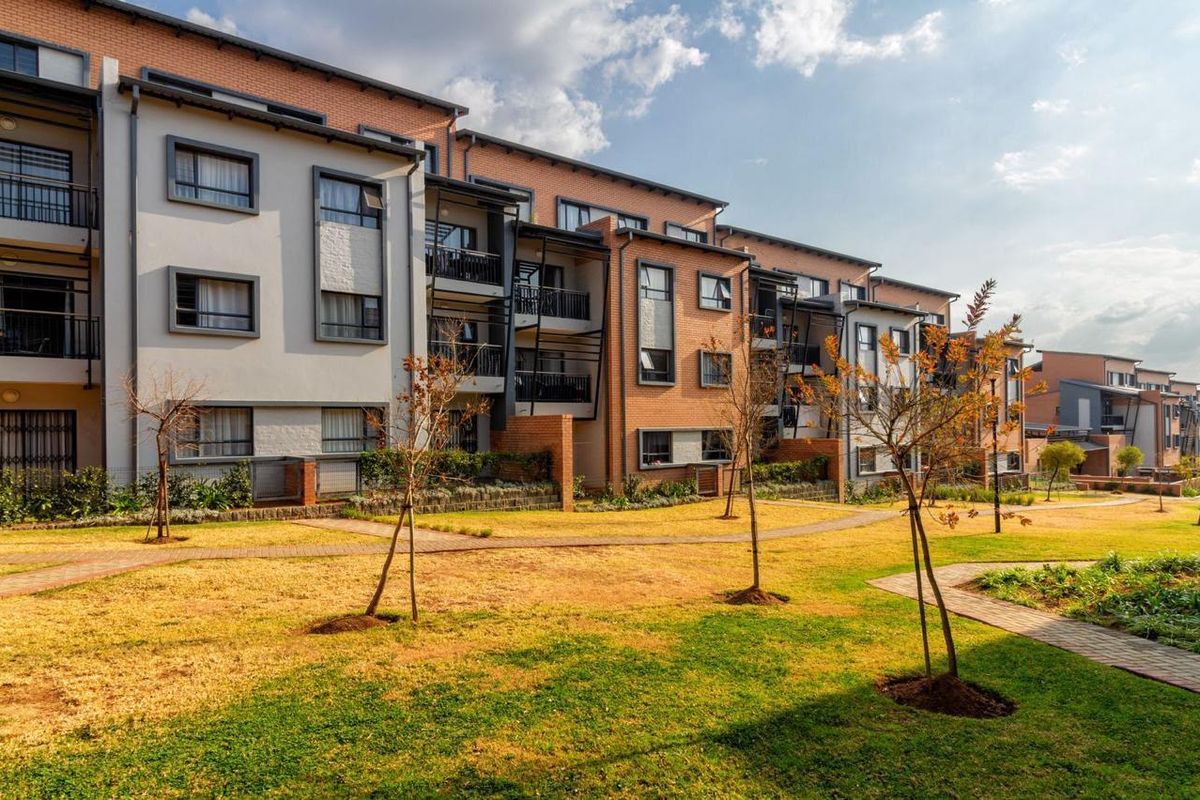 A view of the apartment complex rear facade featuring brick and plaster finishes, private balconies, and well-maintained communal garden areas with paved pathways.