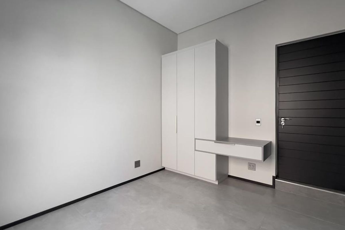 A modern, minimalist bedroom featuring neutral tiled flooring, a sleek built-in wardrobe with an integrated vanity, and a contemporary dark-toned internal door.