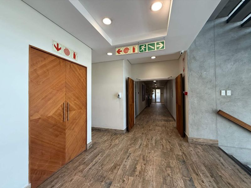A modern, well-maintained office hallway featuring wood-look tile flooring, recessed lighting, and a striking exposed concrete wall accent.