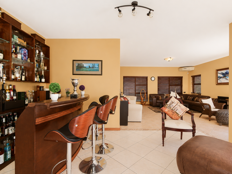 A spacious open-plan living area featuring a dedicated built-in wooden bar with glass shelving, tiled floors, and integrated air conditioning.