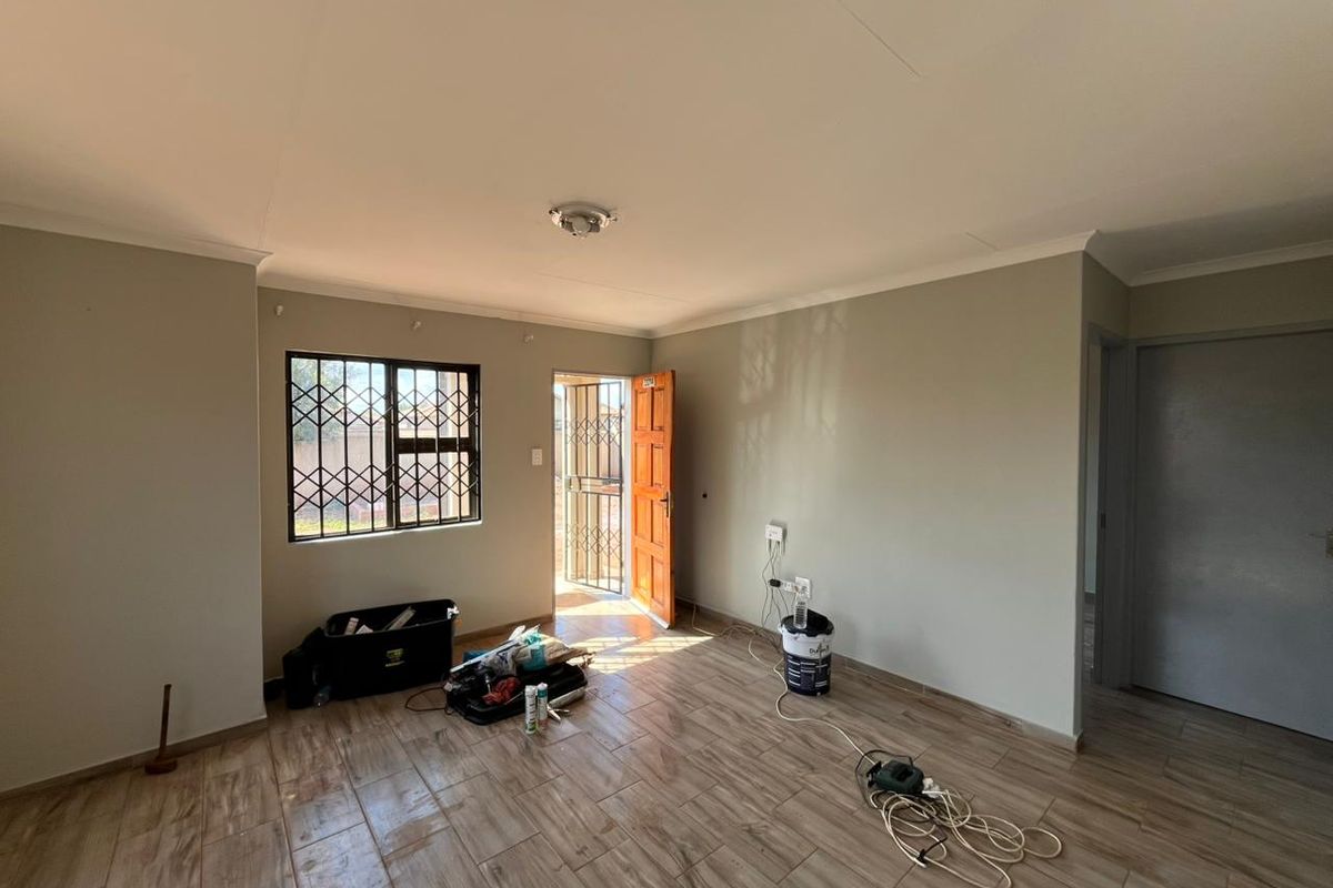 A bright, empty living room featuring wood-look tiled flooring, security bars on the window, and a security gate at the entrance.
