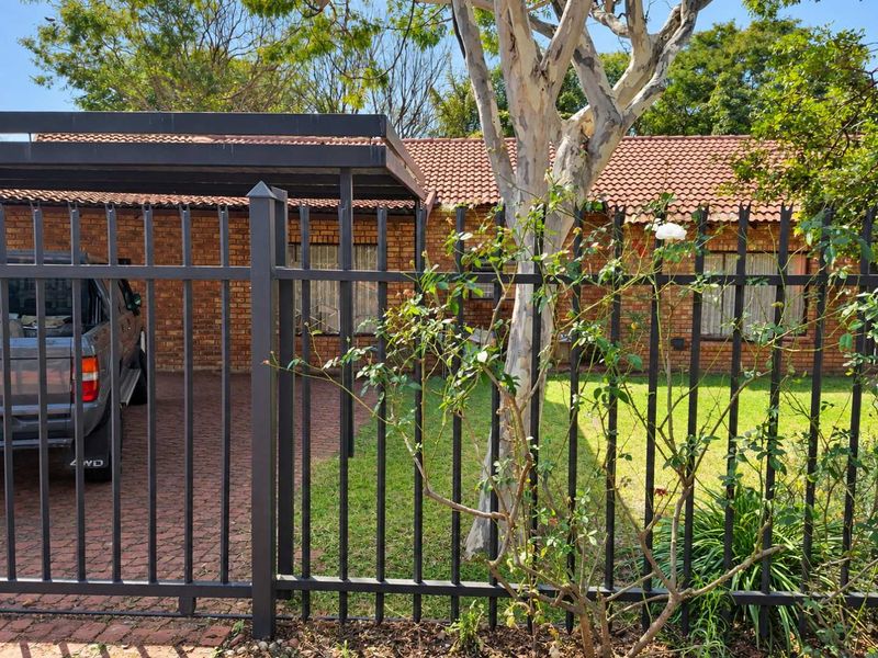 Front view of a brick house featuring a tiled roof, paved driveway, and a covered carport, secured by metal fencing.