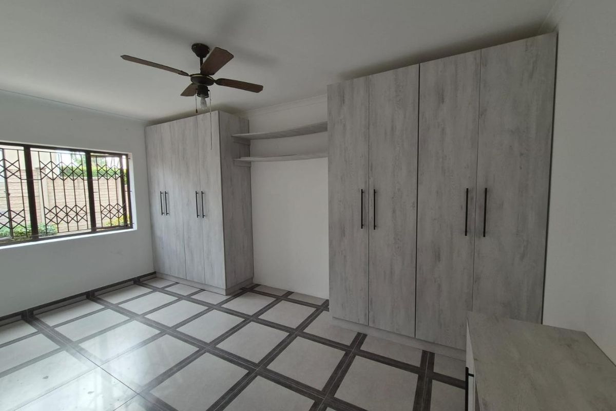 A bright, empty bedroom featuring modern light-wood built-in cupboards, tiled flooring, and a ceiling fan, with security bars on the window.
