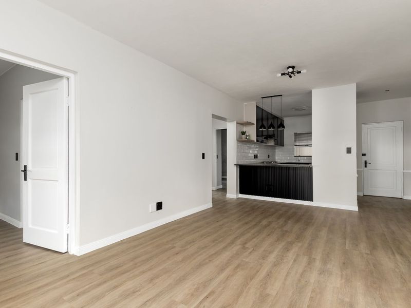 A bright, open-plan living area featuring modern laminate flooring, neutral walls, and a sleek, contemporary kitchen with dark cabinetry and subway tile backsplash.