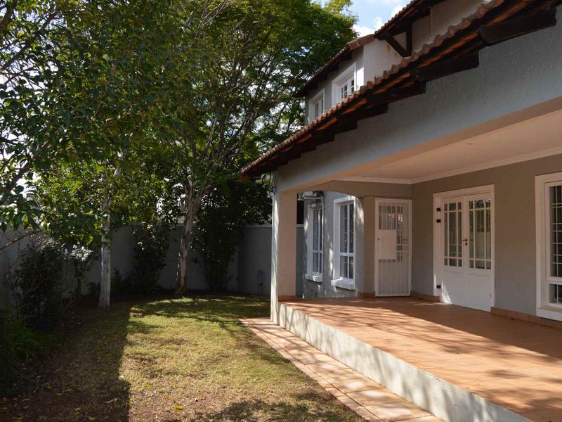 A well-maintained rear exterior view featuring a covered tiled patio, mature garden trees, and white-framed french doors leading into the home.