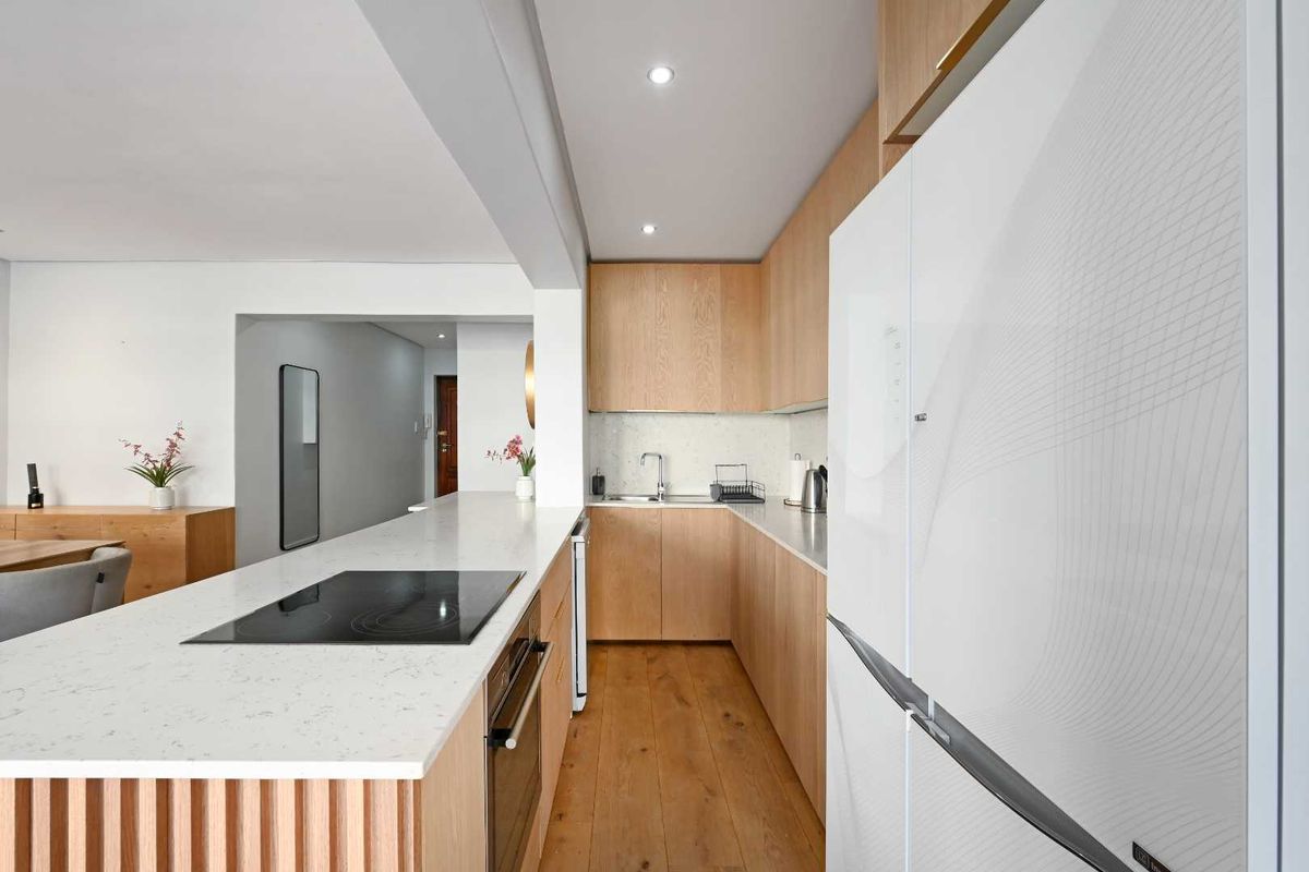 Modern kitchen featuring light wood cabinetry, stone countertops, integrated appliances, and hardwood flooring, seamlessly flowing into the adjacent living area.