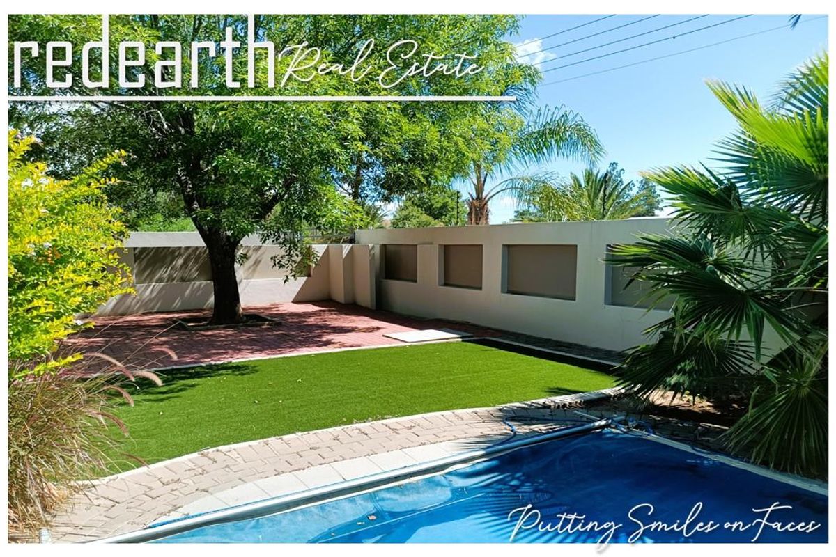 A well-maintained rear garden featuring a swimming pool with a cover, paved patio area, artificial lawn, and a solid perimeter boundary wall.