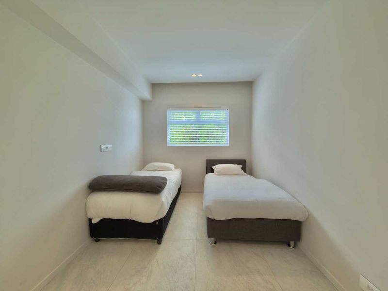 A clean and minimalist bedroom featuring neutral tiled flooring, recessed ceiling lighting, and a window fitted with venetian blinds.