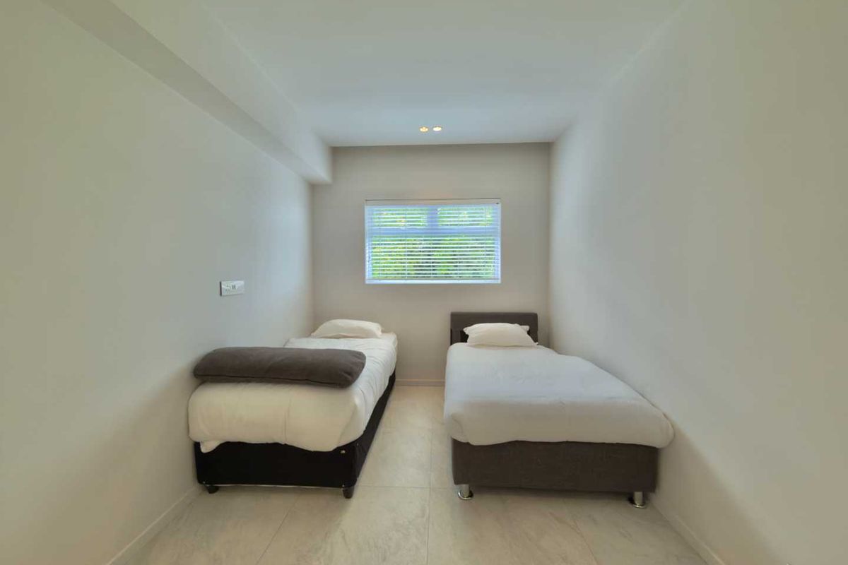 A clean and minimalist bedroom featuring neutral tiled flooring, recessed ceiling lighting, and a window fitted with venetian blinds.
