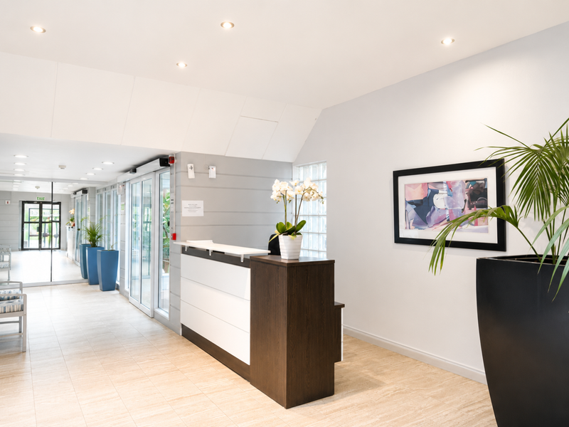 A bright and modern apartment building entrance lobby featuring a reception desk, tiled flooring, and large glass sliding doors leading to the exterior.