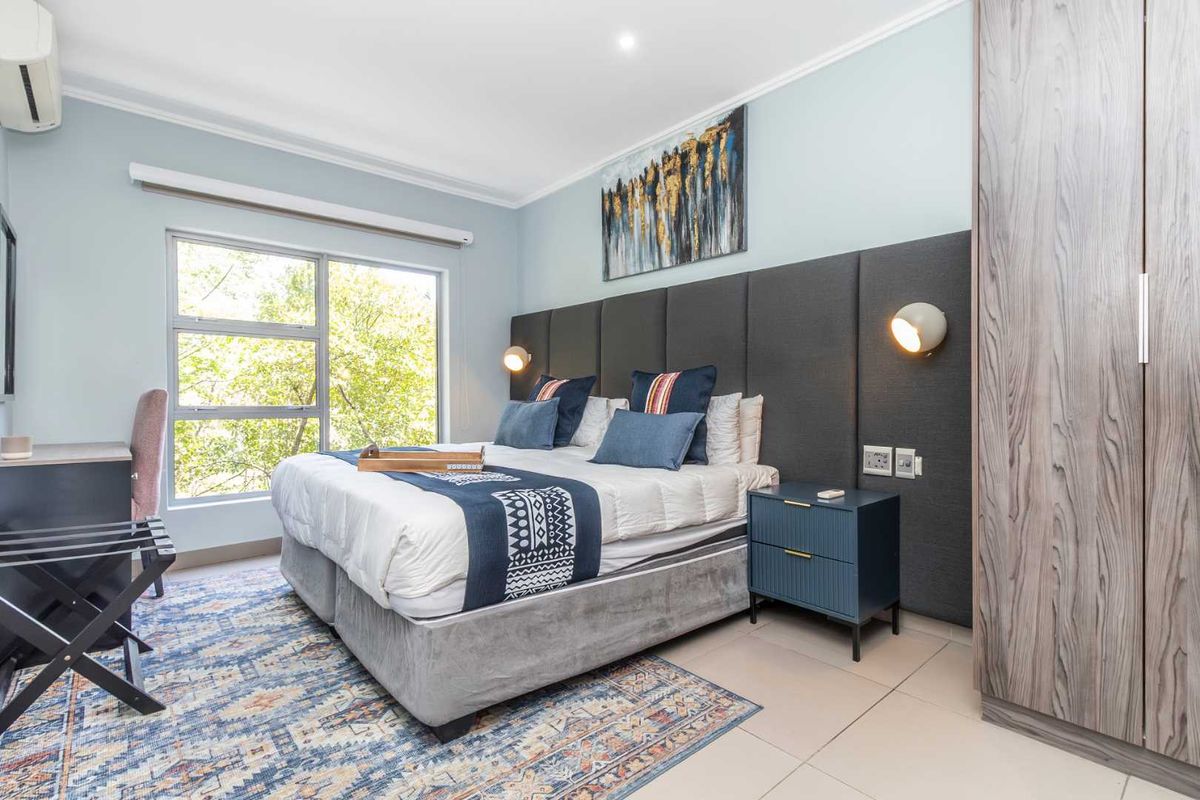 A well-maintained bedroom featuring a large built-in wardrobe, upholstered headboard, wall-mounted lighting, and tiled flooring with a pleasant view through the window.