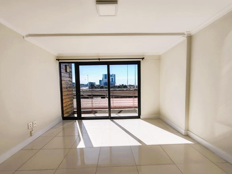 A bright, empty living area featuring neutral tiled flooring and a large sliding glass door providing access to a balcony with urban views.