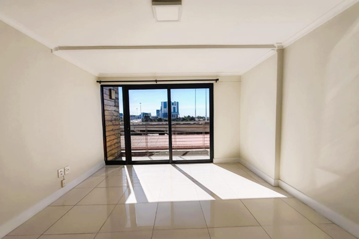 A bright, empty living area featuring neutral tiled flooring and a large sliding glass door providing access to a balcony with urban views.