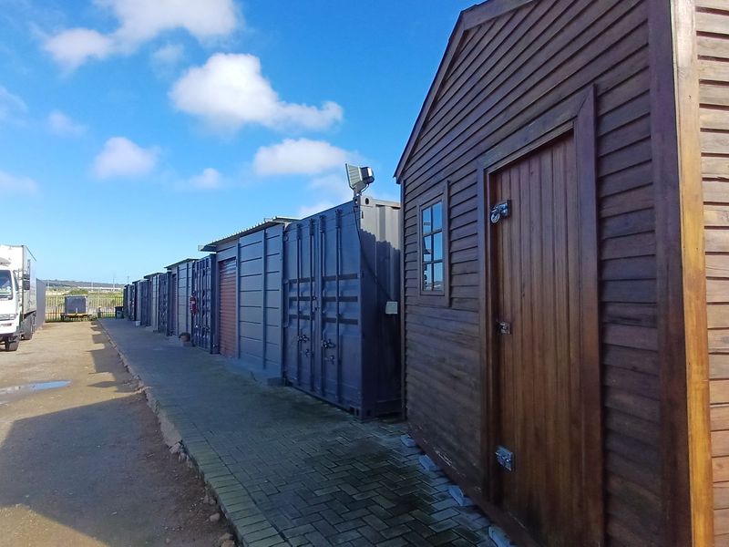 A row of industrial shipping containers and a timber storage shed situated along a paved walkway in an industrial yard setting.