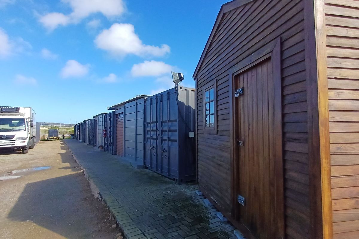 A row of industrial shipping containers and a timber storage shed situated along a paved walkway in an industrial yard setting.