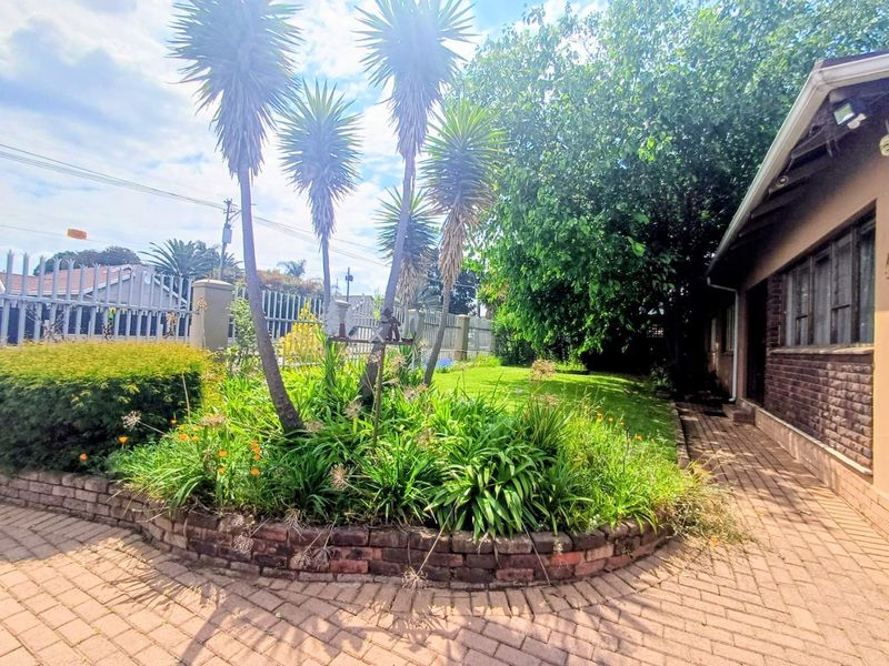 A residential garden area featuring a brick paved path, a raised brick planter bed, mature trees, and metal perimeter fencing.