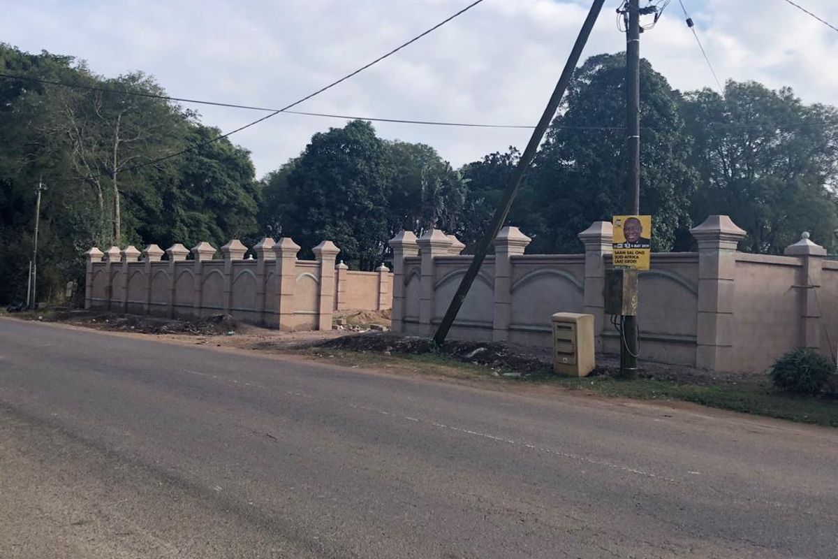 Street view of a residential property featuring a substantial perimeter wall with decorative concrete pillars along the roadside.