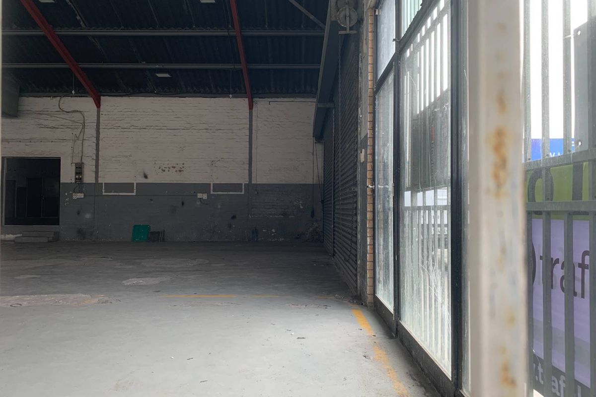 Spacious industrial retail unit featuring high ceilings with exposed steel trusses, concrete flooring, and a large glass storefront with roller shutter access.