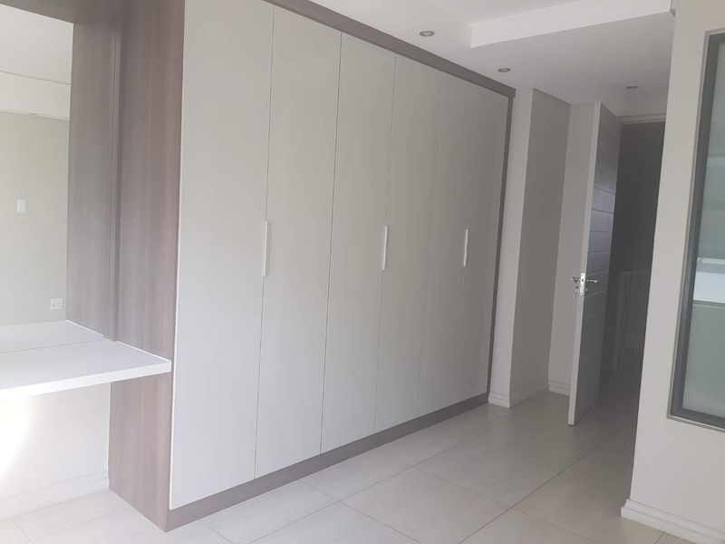 A clean, modern bedroom featuring extensive built-in cupboards, a matching vanity area, neutral tiled flooring, and recessed ceiling lighting.