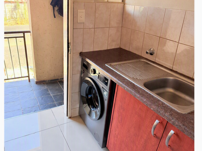 A functional laundry area featuring a stainless steel sink, laminate countertop, tiled backsplash, and built-in cabinetry with direct access to an outdoor balcony.