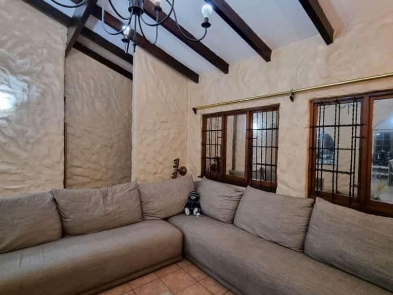 Cozy living room featuring rustic textured walls, exposed timber ceiling beams, tiled flooring, and large timber-framed windows with security bars.