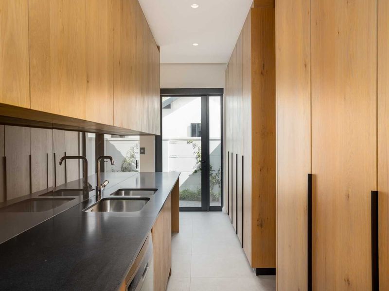 A modern, minimalist scullery featuring extensive timber cabinetry, dark stone countertops, a stainless steel sink, and a mirrored splashback with direct outdoor access.