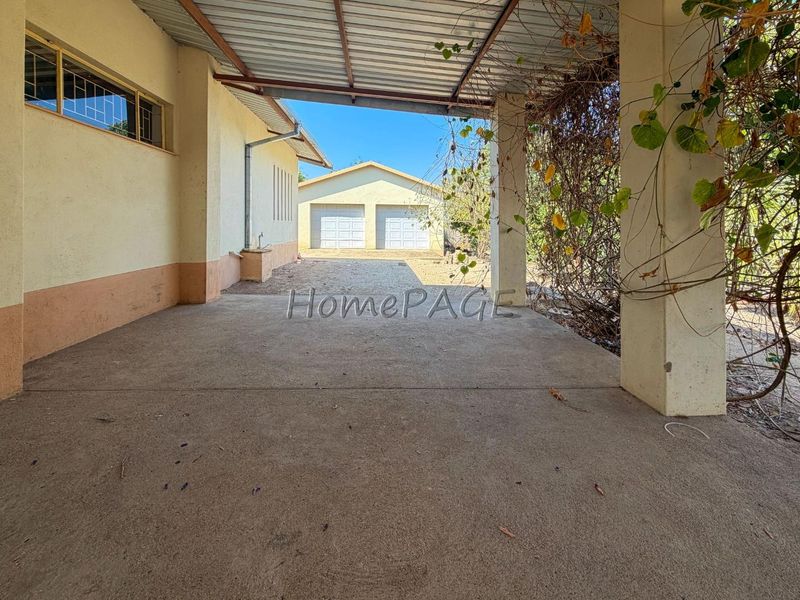 A covered outdoor patio area featuring concrete flooring and support pillars, leading towards a detached double garage in the rear yard.