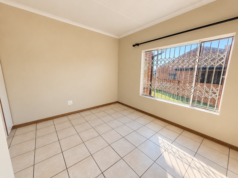 A bright, empty bedroom featuring neutral tiled flooring, clean walls, and a window fitted with a security gate.