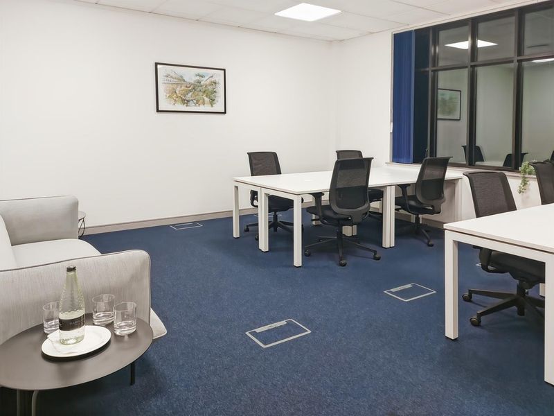 A functional office space featuring blue carpeting, recessed floor power outlets, and a large window with a dark frame.