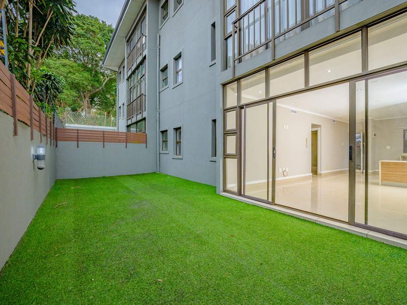 A private rear garden area featuring artificial turf, a secure boundary wall, and large sliding glass doors providing access to the apartment interior.